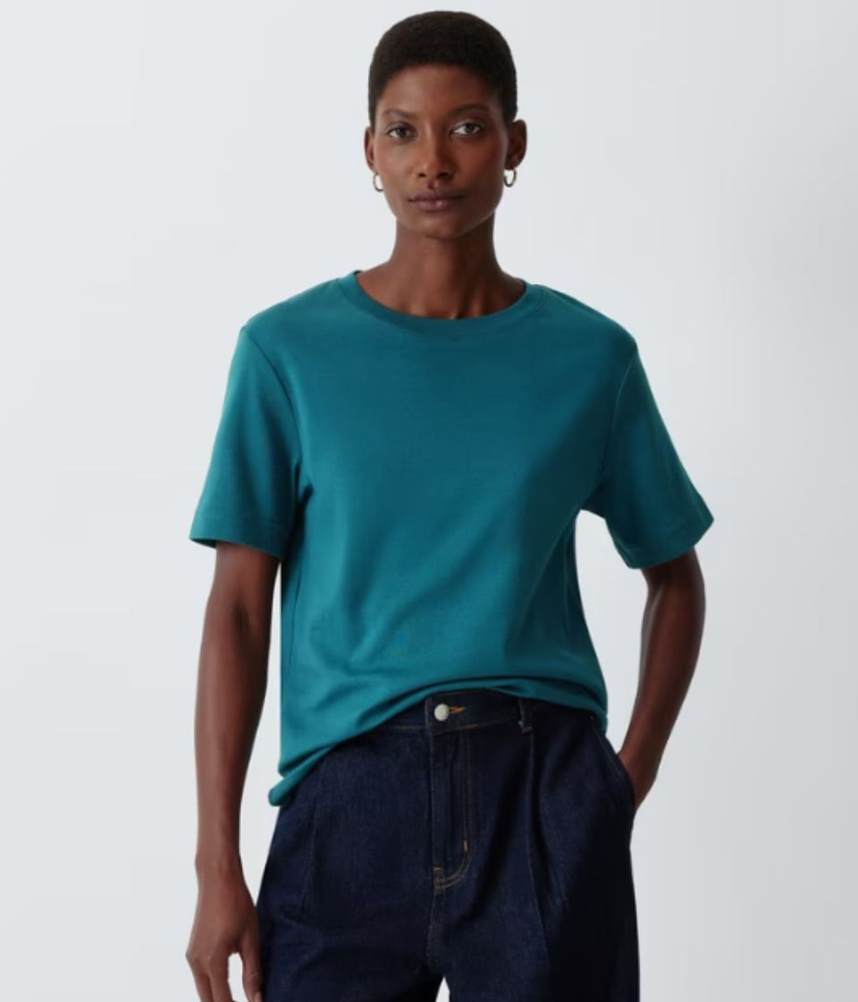 A model wearing a teal short-sleeve crewneck t-shirt and dark blue jeans, standing against a light gray studio background.