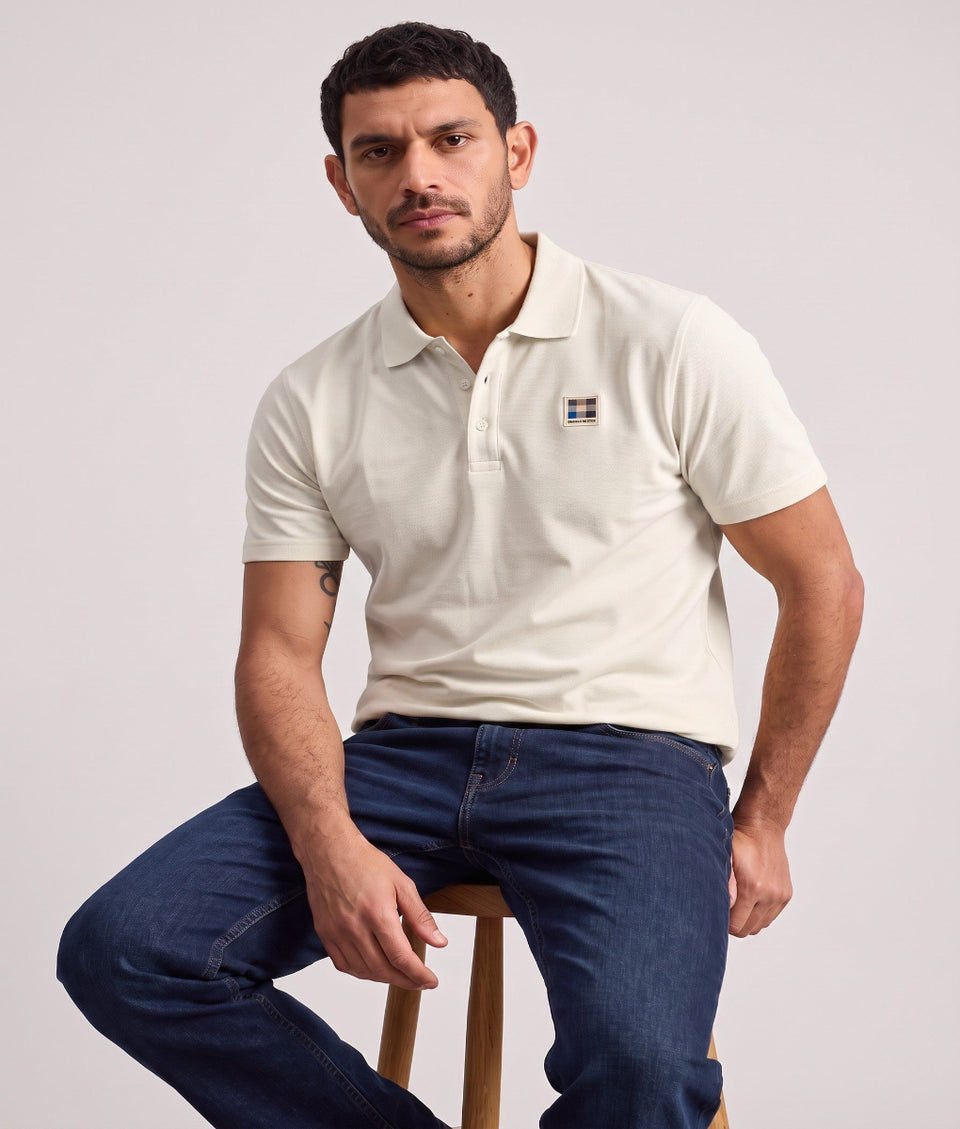 A man wearing a cream-colored short-sleeve polo shirt with a small embroidered logo on the chest, paired with dark blue jeans, sitting on a wooden stool against a light gray background.