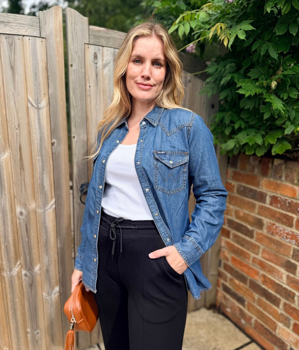 A woman stands outdoors in front of a wooden fence and brick wall, wearing a blue denim button-up shirt over a white top with black drawstring pants, and holding a brown leather clutch.