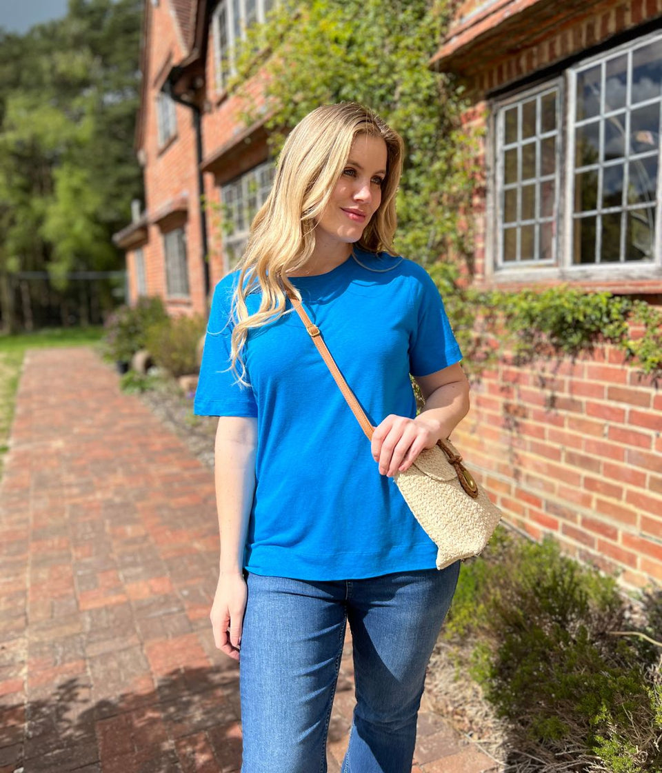 Woman in a blue shirt and jeans holding a beige bag, standing on a brick path with a house in the background.