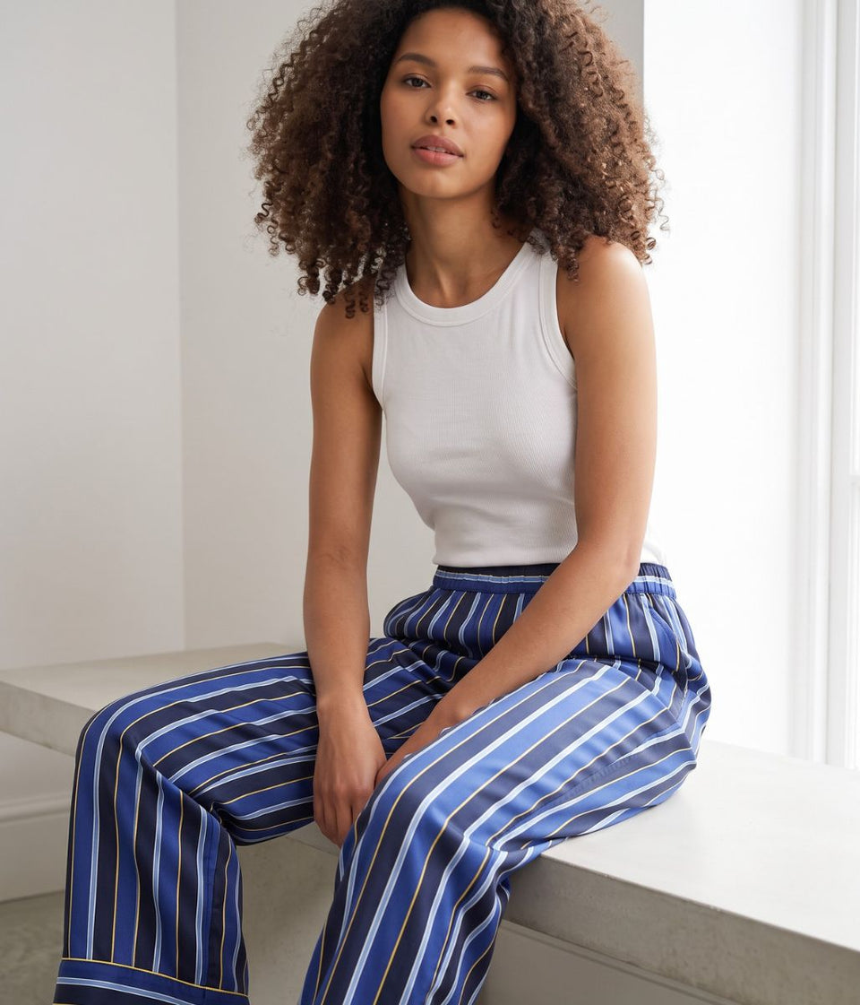A woman with curly brown hair sits on a light-colored stone bench wearing a white ribbed tank top and blue wide-leg trousers with vertical white and thin gold stripes.
