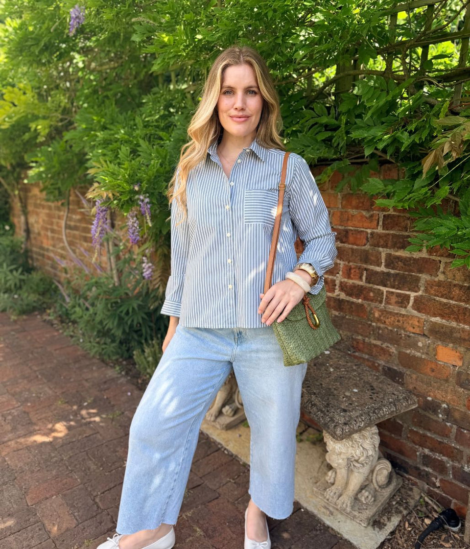 A woman stands outdoors in a garden setting wearing a light blue and white striped button-down shirt, light-wash cropped jeans, and white sneakers. She is carrying a green woven crossbody bag with a brown strap.