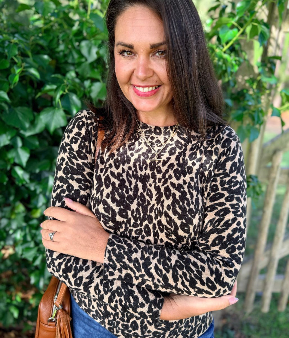 A woman wearing a beige and black leopard print long-sleeve top, standing outdoors with her arms crossed and smiling.