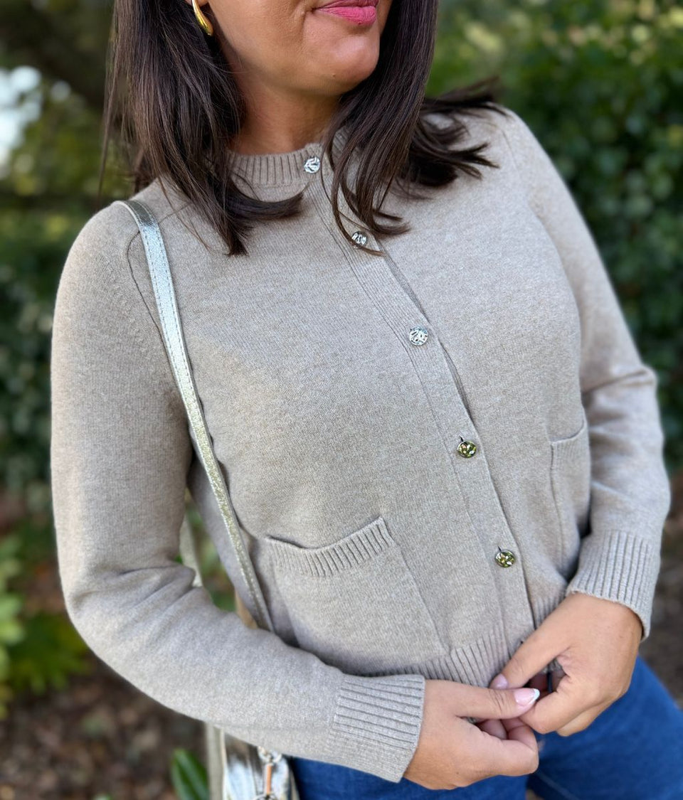 A woman wearing a light taupe knit cardigan sweater with decorative metallic buttons, paired with blue jeans and a silver shoulder bag, set against a blurred green outdoor background.