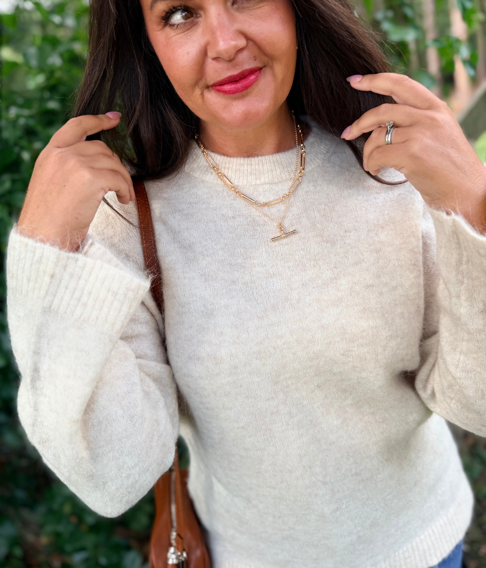 A woman wearing a fuzzy off-white crewneck sweater, styled with layered gold T bar necklaces, a brown shoulder bag, and blue jeans, set against a soft-focus green outdoor background.