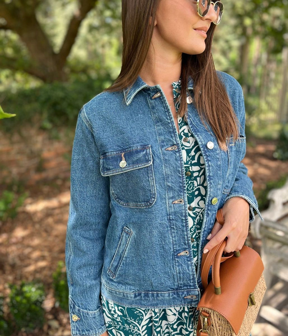 Woman wearing a denim jacket and holding a brown leather bag outdoors.