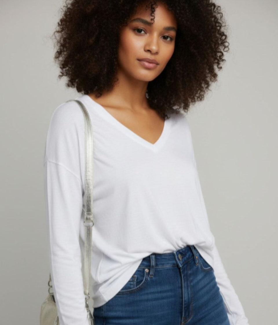 A woman with curly dark hair wearing a white long-sleeve V-neck top and blue jeans, with a light-colored shoulder bag, against a plain light gray background.